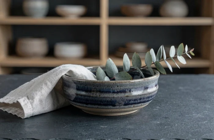 Gros plan d'un couvert artisanal avec bol en céramique émaillée indigo, serviette en lin texturée et branche d'eucalyptus séché sur ardoise